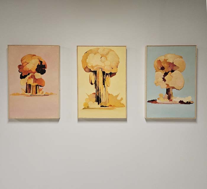 Three paintings of stylized nuclear bomb tests, in soft colors of red, yellow, and blue.