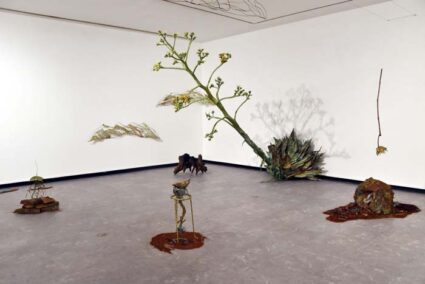 Installation view of multi-media artworks resembling natural forms, including a large sculpture of an blooming century plant vaulting out at a slant from the wall into the gallery.