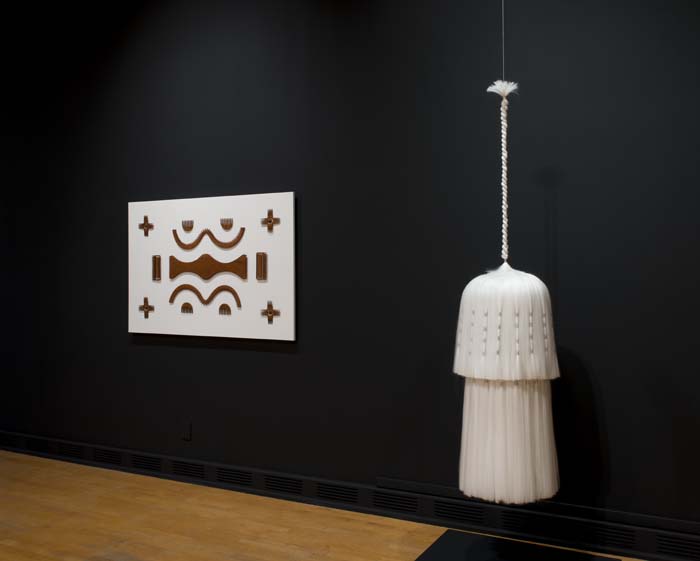 Installation view of two works, on the left a beautifully crafted puzzle-like piece made of African mahogany combs, and on the right a sumptuous chandelier-like suspended work made of white Kanekalon hair extensions.