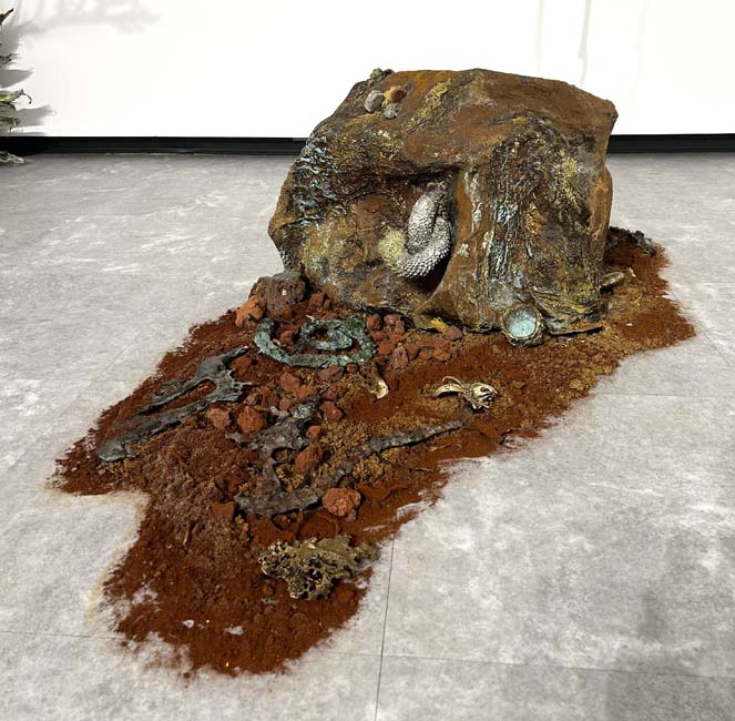 Sculpture of a naturalistic rock strewn with cast bronze spills laid out on rust red sand.