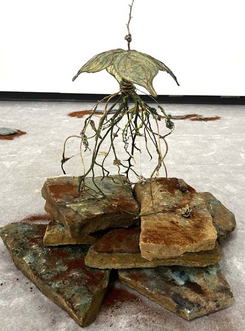 Sculpture of cast bronze made to appear like the roots of a plant floating over a stack of rocks dusted with red volcanic sand.