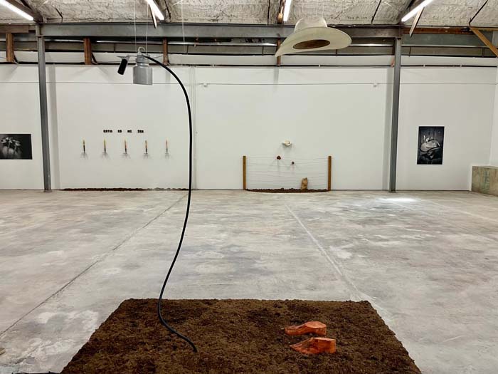 Installation view of José Villalobos: Fuertes y Firma, with a sculptural installation in the foreground consisting of a square of soil with a pair of clay feet, a suspended white hat and a pesticide canister.