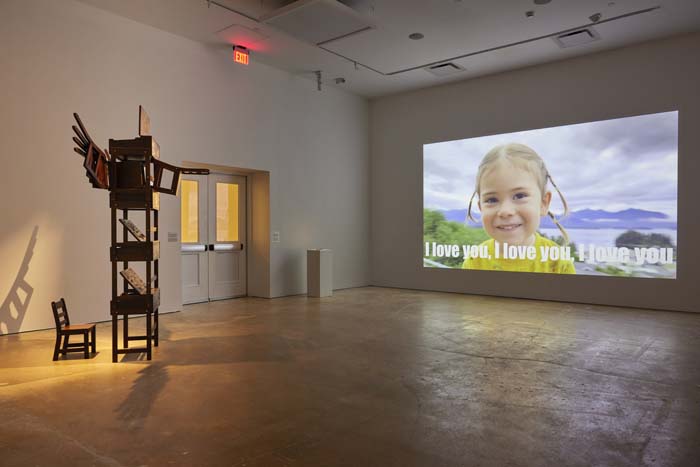 Installation view with an eight-foot-tall wooden sculpture resembling a totem pole of children's desks at left, and a large video projection of a smiling child in a yellow shirt with the words "I love you" in subtitles.