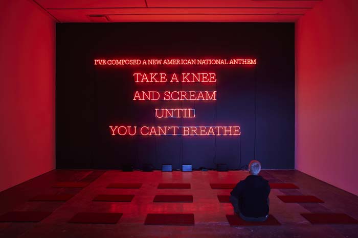 An installation with the words “I’ve composed a new American national anthem / Take a knee / and scream / until / you can’t breathe,” in all-capped red neon lettering against a black wall.