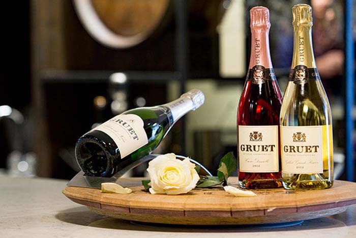 Three bottles of wine by Gruet Winery, with one bottle tilted on its side, set on a wooden board with a white rose.