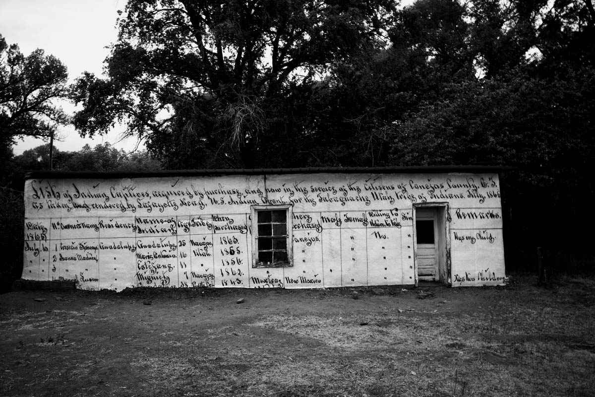 Black and white photograph of a low house wheatpasted with a handwritten list of names of Native enslaved people.