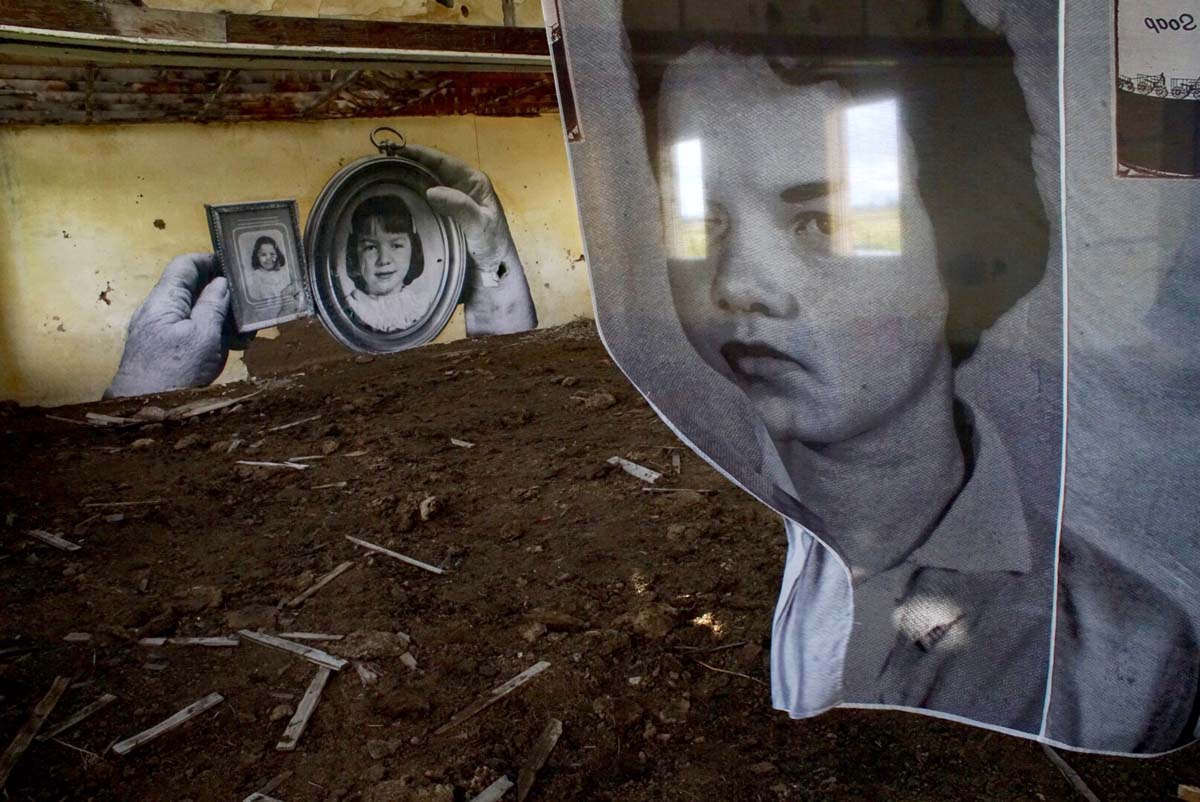 Interior of an abandoned building with a black and white photograph of a woman printed on fabric hanging in the space, and two other photographs pasted on the wall in the background.