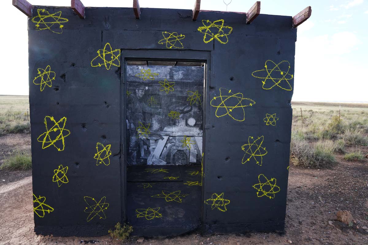A small outbuilding painted black with yellow radioactivity symbols, with a black and white photograph of a uranium miner wheatpasted in the window.