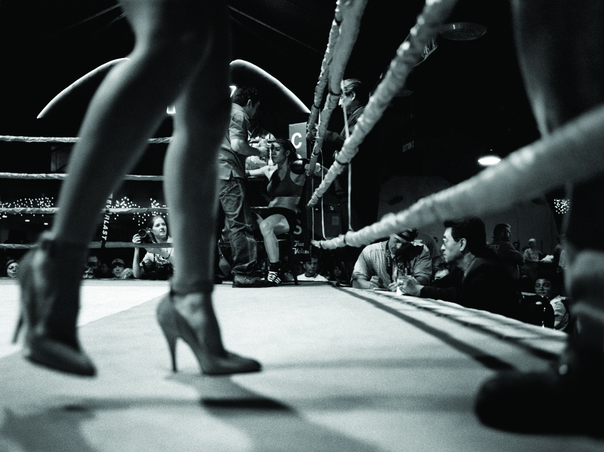 Black and white photograph by Delilah Montoya showing blurred, out-of-focus image of woman’s legs in high heels walking in a boxing ring in front of a female boxer in the back corner of the ring. Women Boxers: The New Warriors, 2006. 