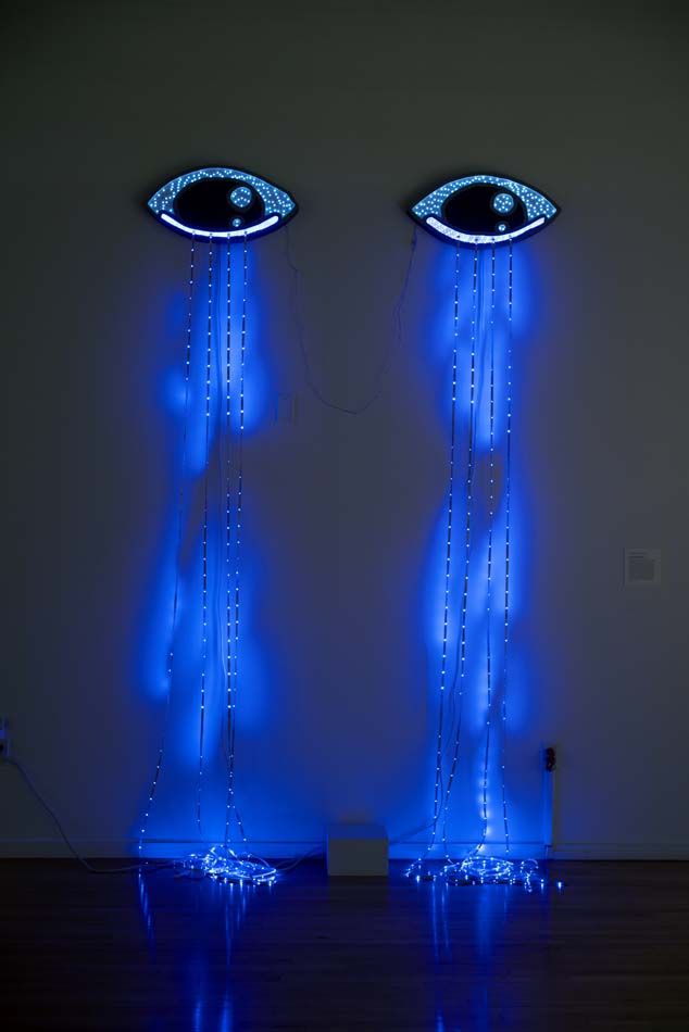 Oversized eyes hanging on a wall with blue LED lights like tears streaming from them down the wall.