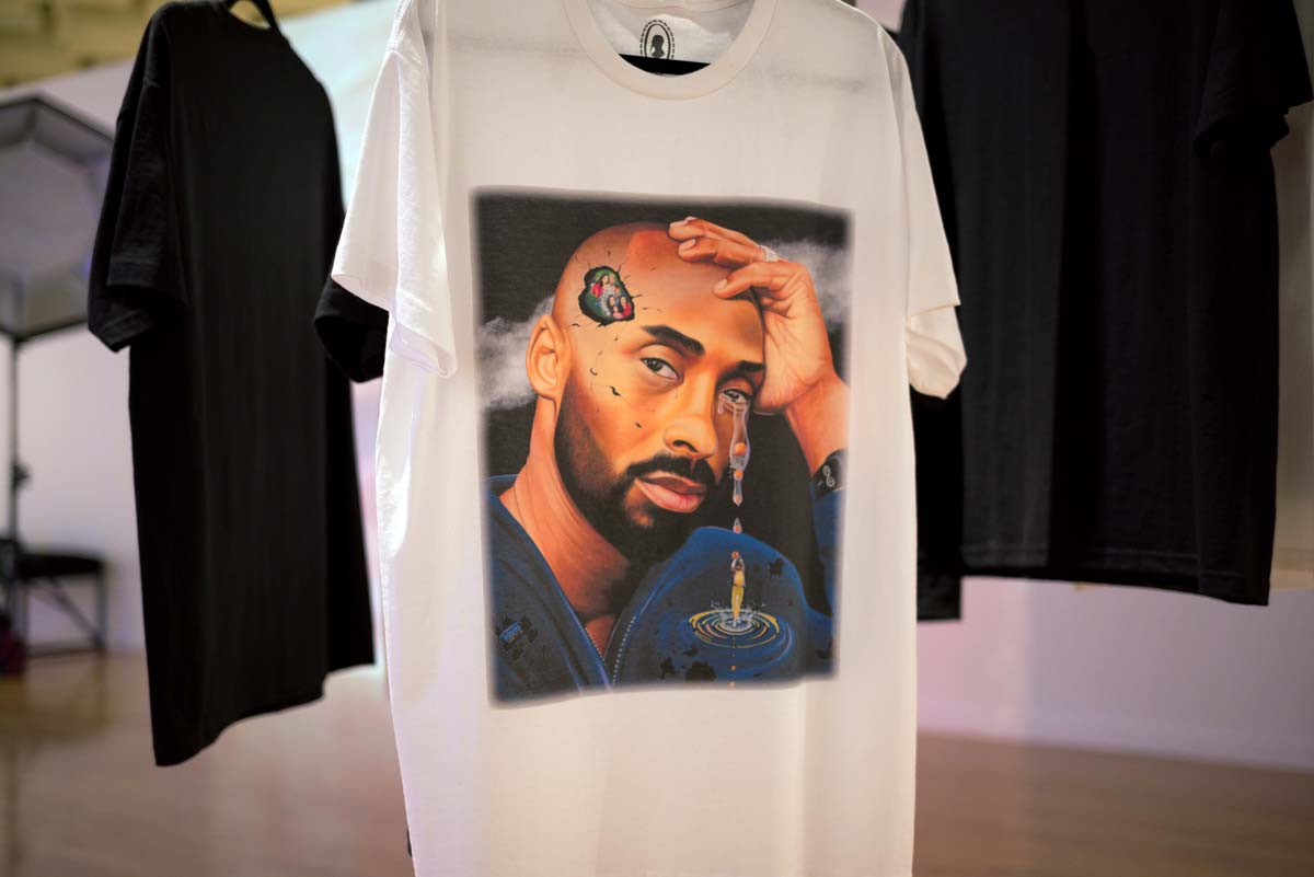White t-shirt on a hanger, with a painting of a Black bearded man's face, tears streaming from his eye, with a hole in the side of his head revealing a family portrait inside.