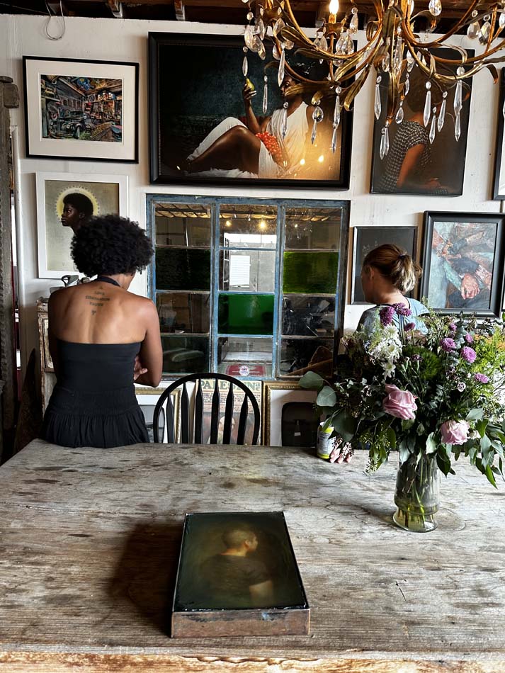 Two people with their backs to the viewer looking at a wall of artwork with a photograph of a person's back lying on a table behind them.