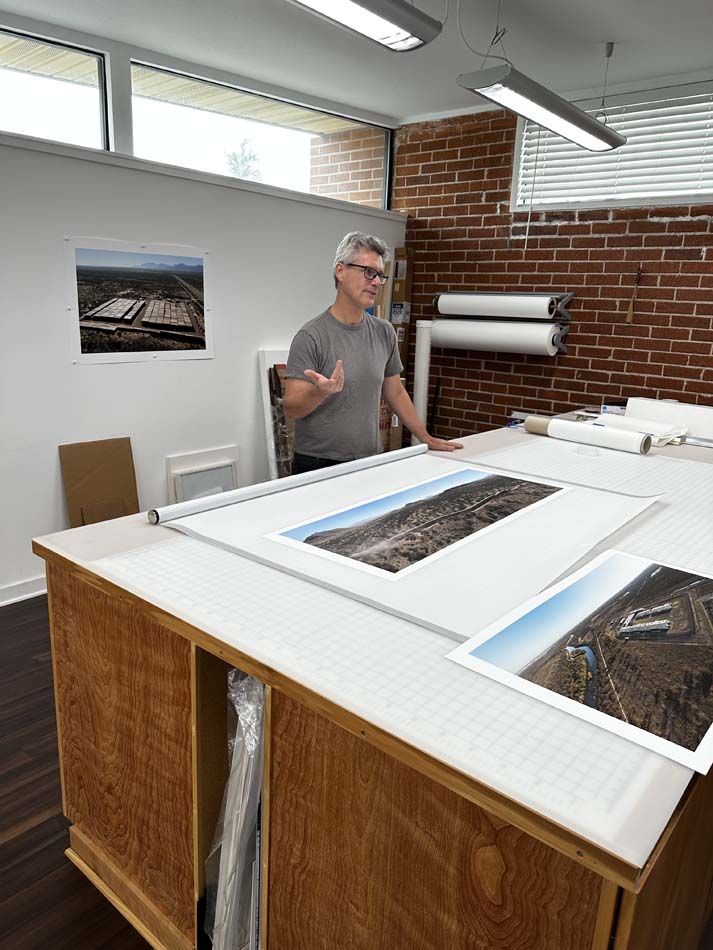A man in a grey t-shirt showing large-scale landscape photographs on a white table.