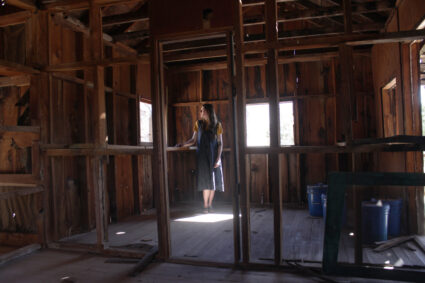 Writer Emma Kemp standing and looking out the window of an abandoned cabin on the grounds of a former religious commune founded by Marie Ogden, Home of Truth in Dry Valley, Utah. Photography by Emily Arntsen.