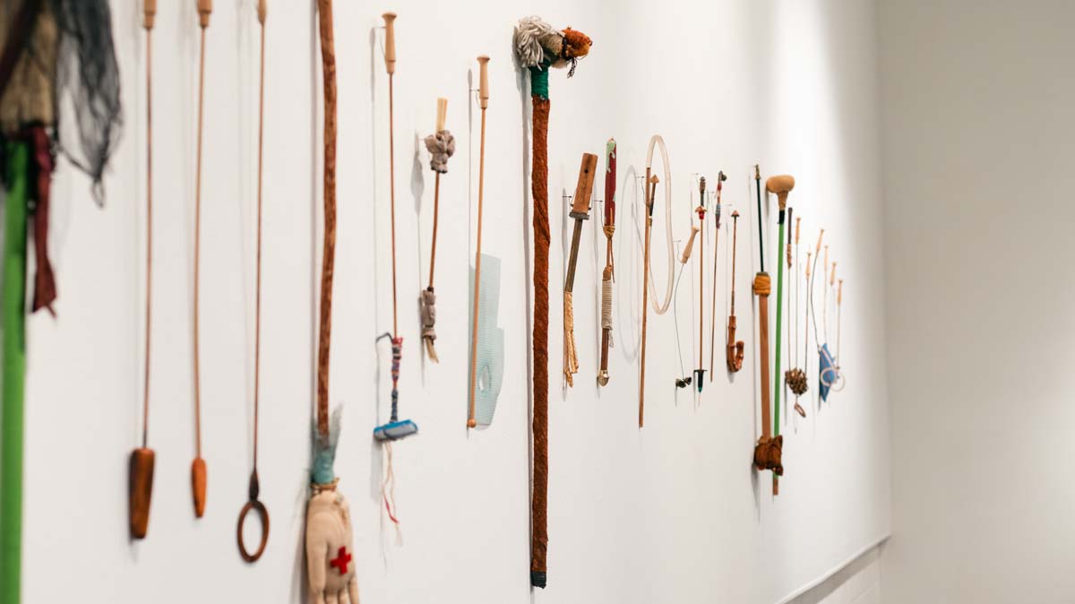 A collection of wands by Jerry Hunt made of everyday objects attached to rods or sticks, hung on a wall.