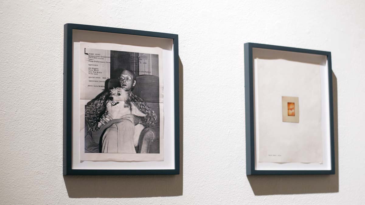Two framed pieces of ephemera on a wall, on the left a flyer for a Jerry Hunt performance with the artist pictured with his dog, and on the right a polaroid that is just out of focus.