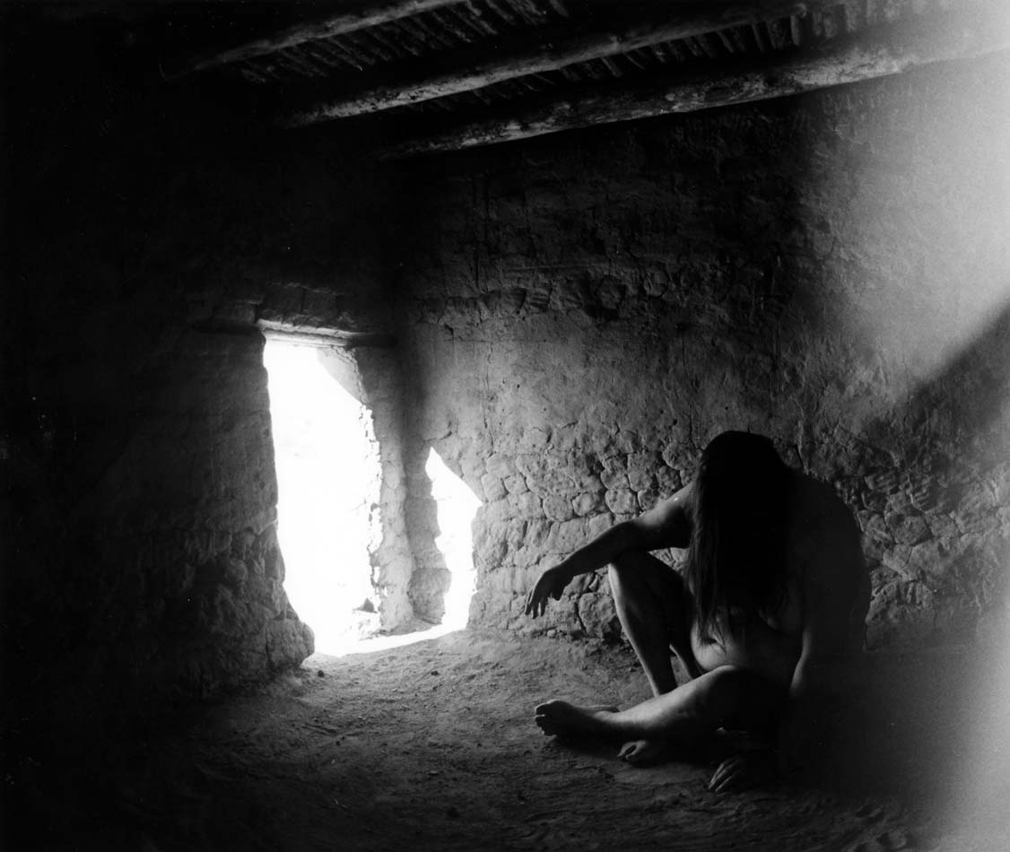 Laura Aguilar posing seated inside a cliff dwelling where it appears her head is hung in despair.