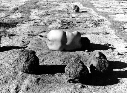 Artist Laura Aguilar lying nude with her back to the camera, in a black and white photograph with similarly shaped boulders.