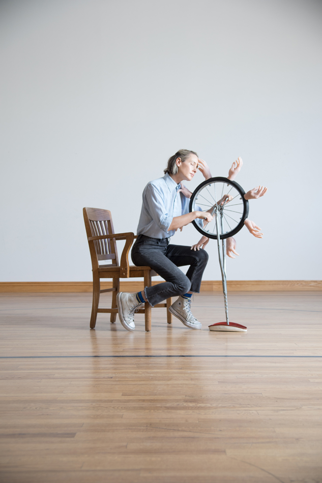 Laura Shill sits at a chair in front of a upside down unicycle containing silicone hands 