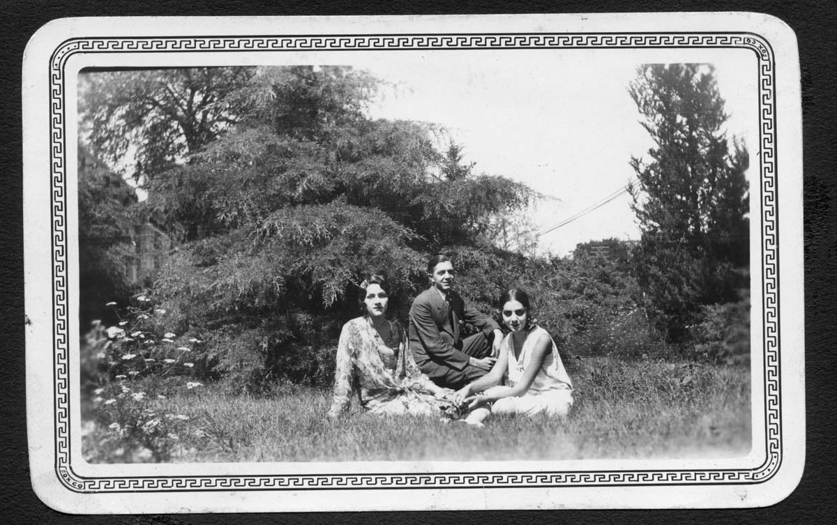 Black and white photograph of a man sitting with his two sisters in a grassy field with trees behind them.