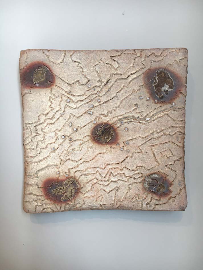 A white glazed ceramic wall piece with five reddish-brown dots placed like the pattern of a die, with meandering, topographical lines throughout.