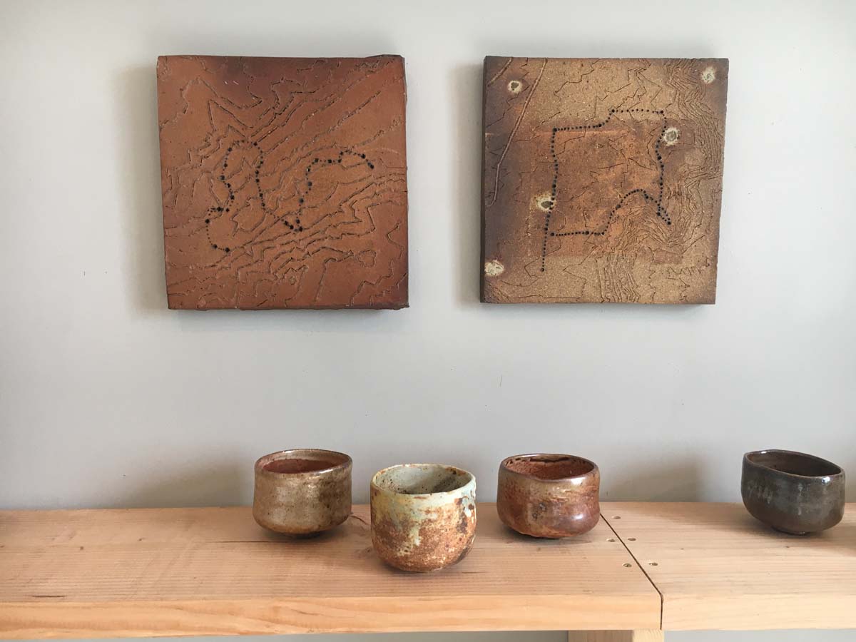 Two nearly square ceramic wall hangings with meandering lines inscribed in them above a wooden shelf with various ceramic tea bowls.