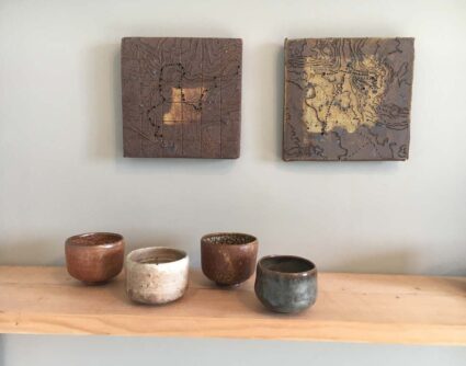 Two nearly square ceramic wall hangings with meandering lines inscribed in them above a wooden shelf with various ceramic tea bowls by Bill Gilbert.