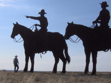 Two twenty-foot tall metal silhouettes of horse mounted cowboys, one pointing off into the distance, with a man standing next to it for scale.