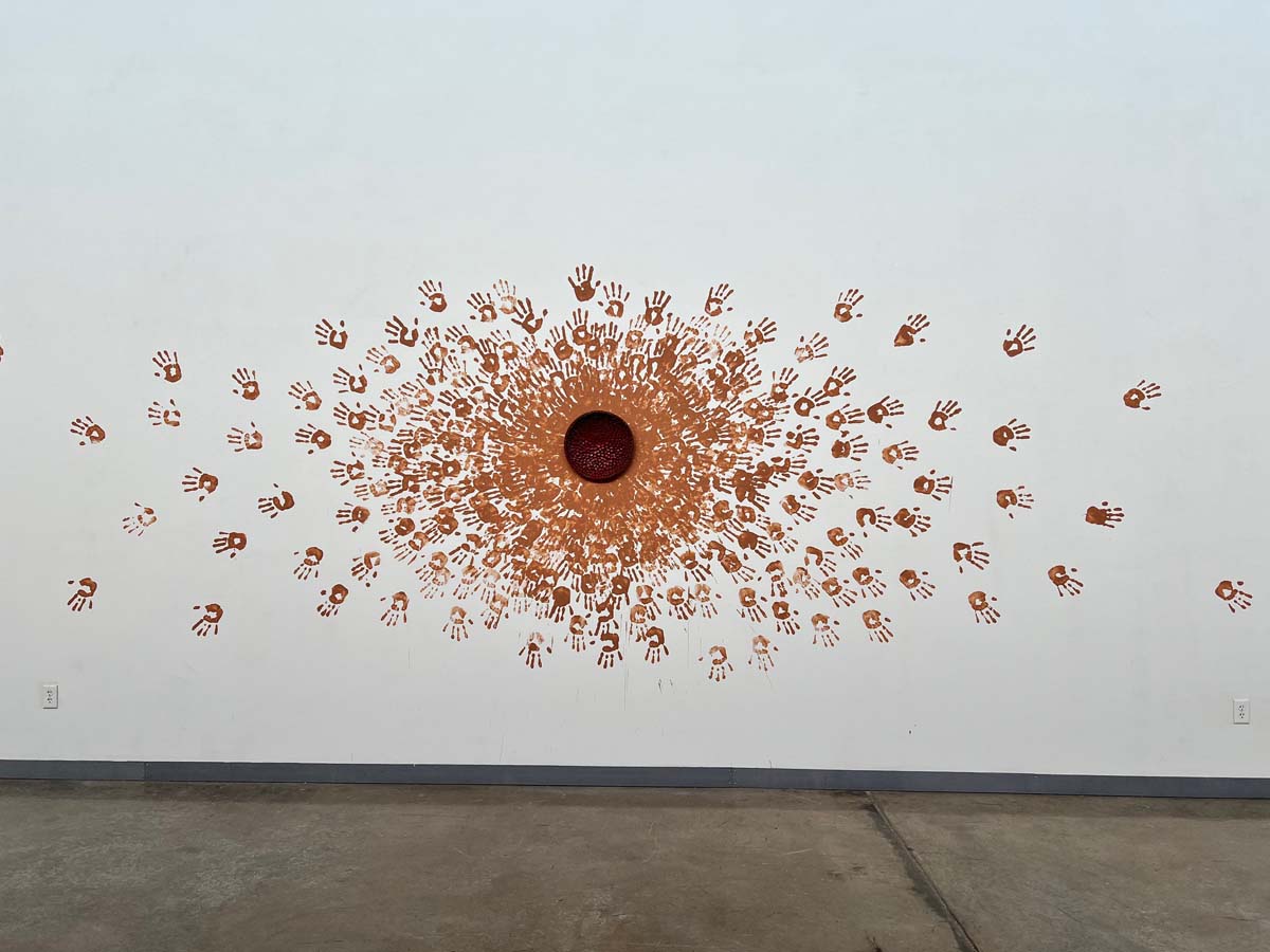 A red-glazed stoneware bowl forms the center of an installation of handprints emanating from it on a wall.