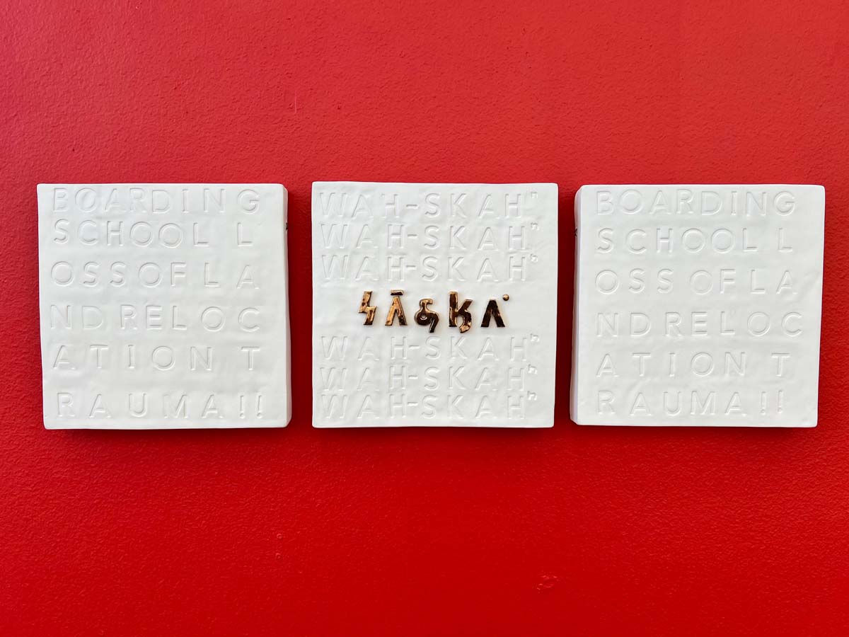 Three ceramic squares with white glaze and the words "boarding school loss of land relocation trauma" imprinted in the two outer panels, and the word "wah-skah" in the central panel in Osage lettering in gold.