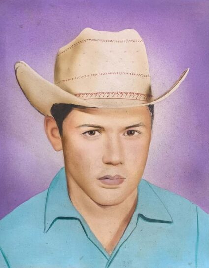 Portrait of a young man wearing a blue collared shirt and a cowboy hat in front of a lavender background.