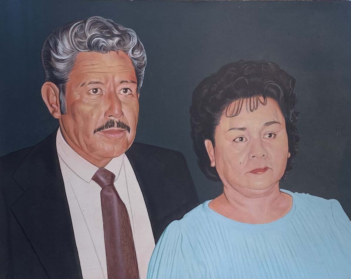 Portrait of an unsmiling Mexican married couple, the man with silver hair and suit and tie, and the woman wearing light blue.