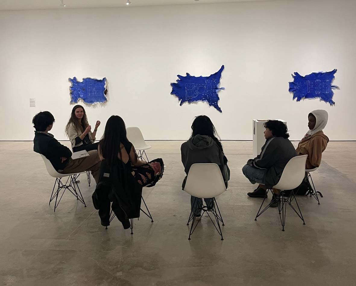SITE Santa Fe's Young Curators meeting in a gallery to discuss art and curatorial methods in Nicholas Galanin's 2023 exhibition at SITE, with a series of blueprint-colored animal hides on the wall behind them.