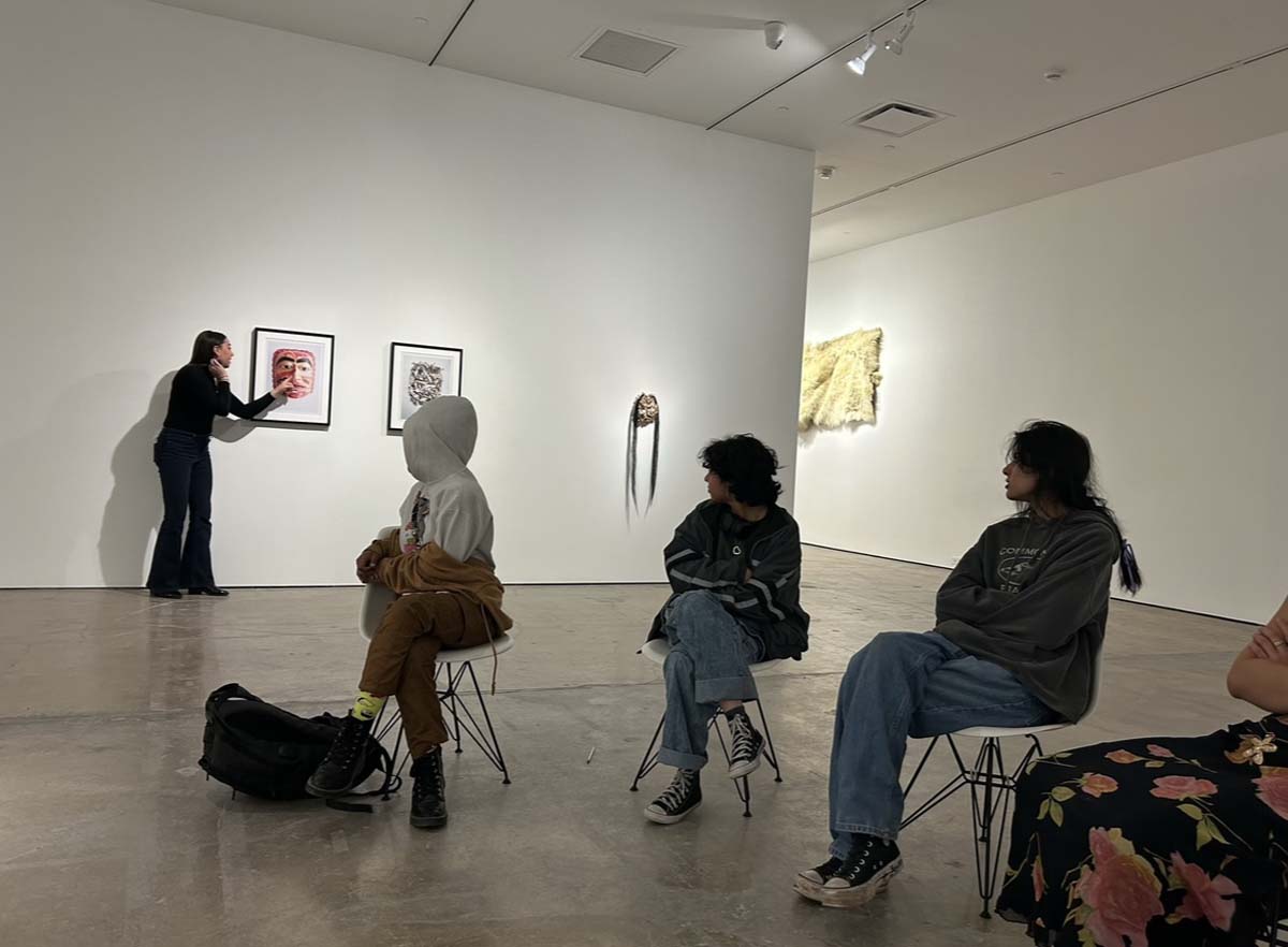 A woman pointing to an artwork of a mask on a wall, with several seated figures looking over their shoulders at her.