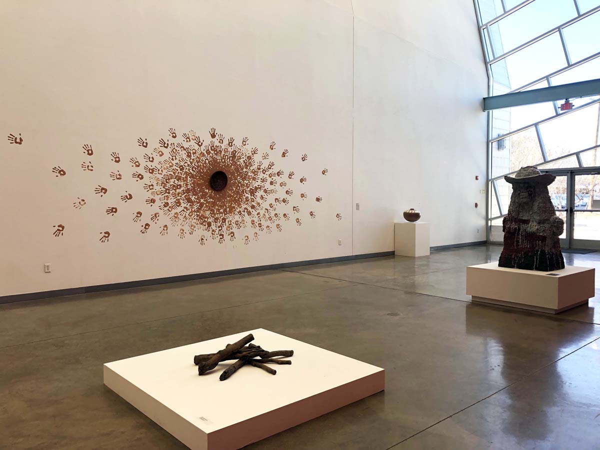 Installation view of Belonging: Contemporary Native Ceramics from the Southern Plains with a large-scale ceramic bust of a woman with a cowboy hat on, a stack of ceramic logs, and a wall installation with handprints radiating out from a central circle.