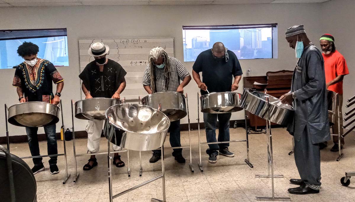 A group of six Black men playing steel pans inside a brightly lit room. 