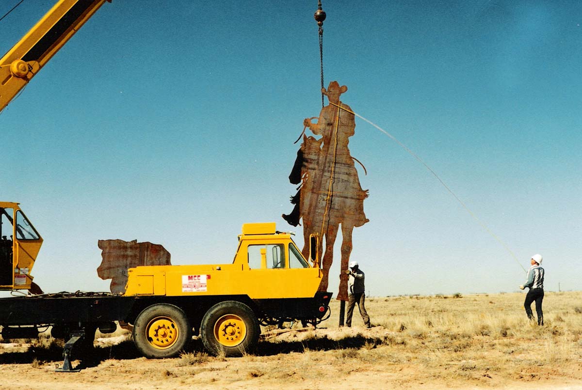A crane lifting a twenty-foot tall metal silhouette of a cowboy on horseback.