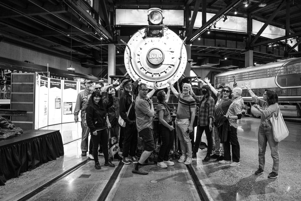 Artists and project participants from the railroad-themed art exhibtion The Other Side of the Tracks pose in front of a historical steam engine.