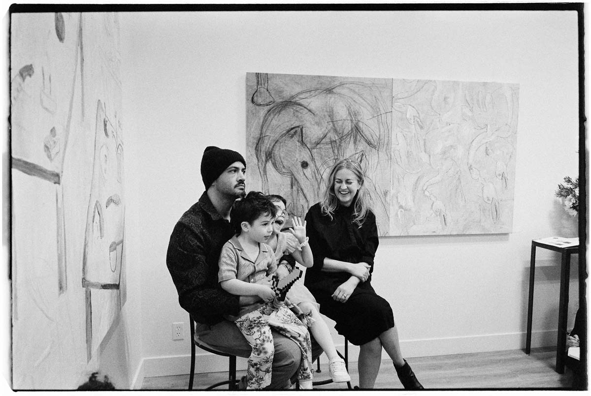 Black and white photograph of the artist Andrew Alba sitting with his children on his lap in a gallery.