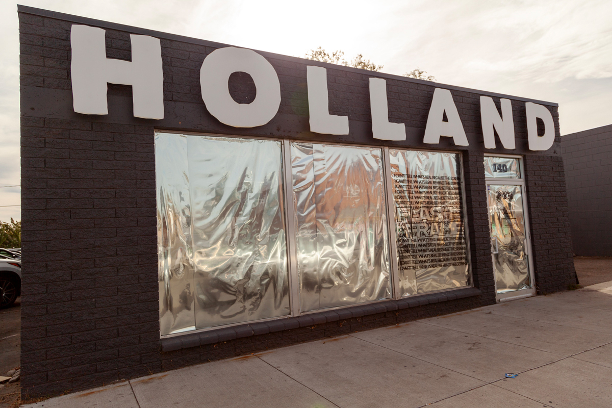 Exterior of a contemporary art gallery with large-scale cutout lettering that reads "HOLLAND." The expansive windows are mysteriously covered with shimmering silver sheets.
