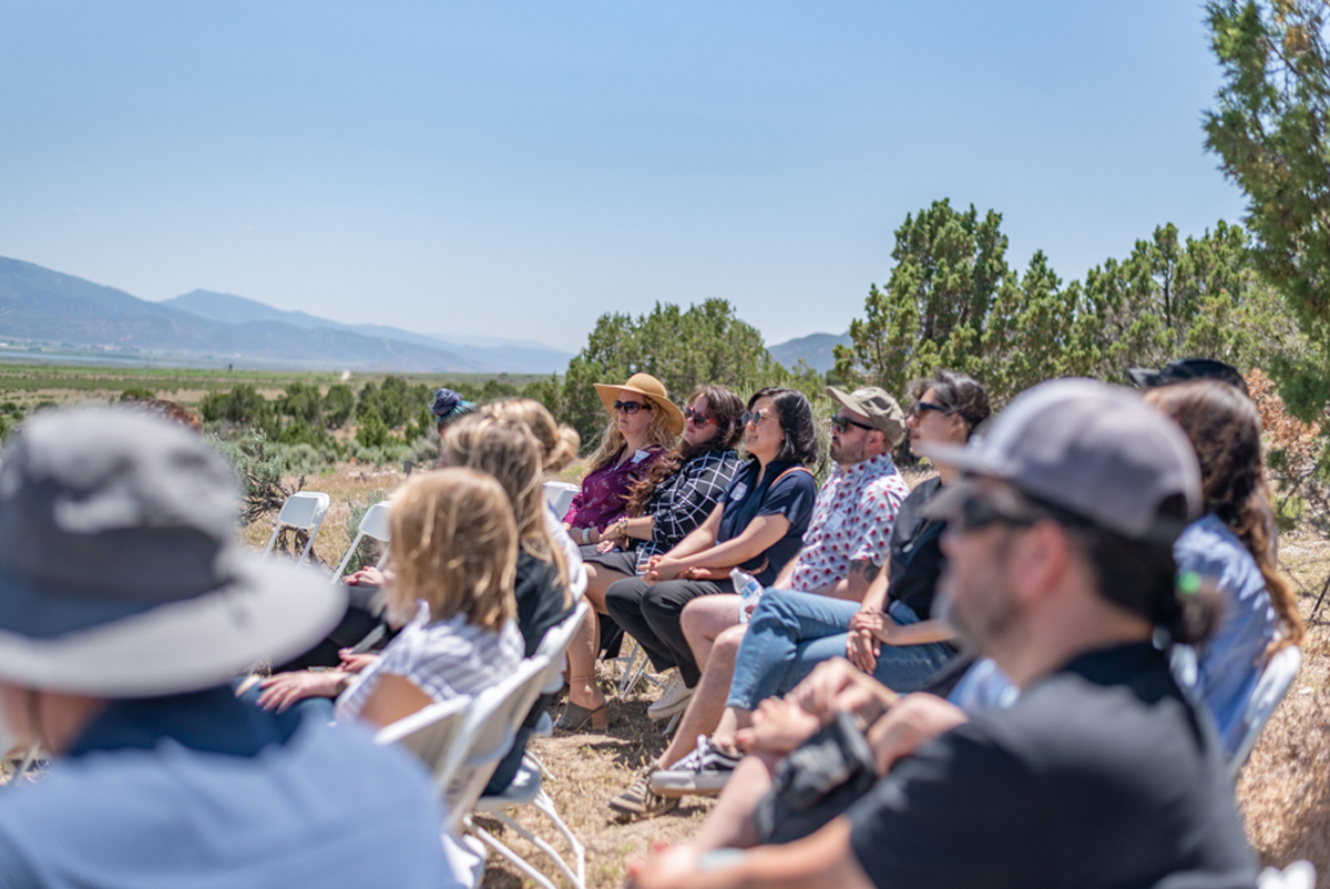 People pack several rows of white folding chairs, foregrounding a Southwestern vista in rural Utah.