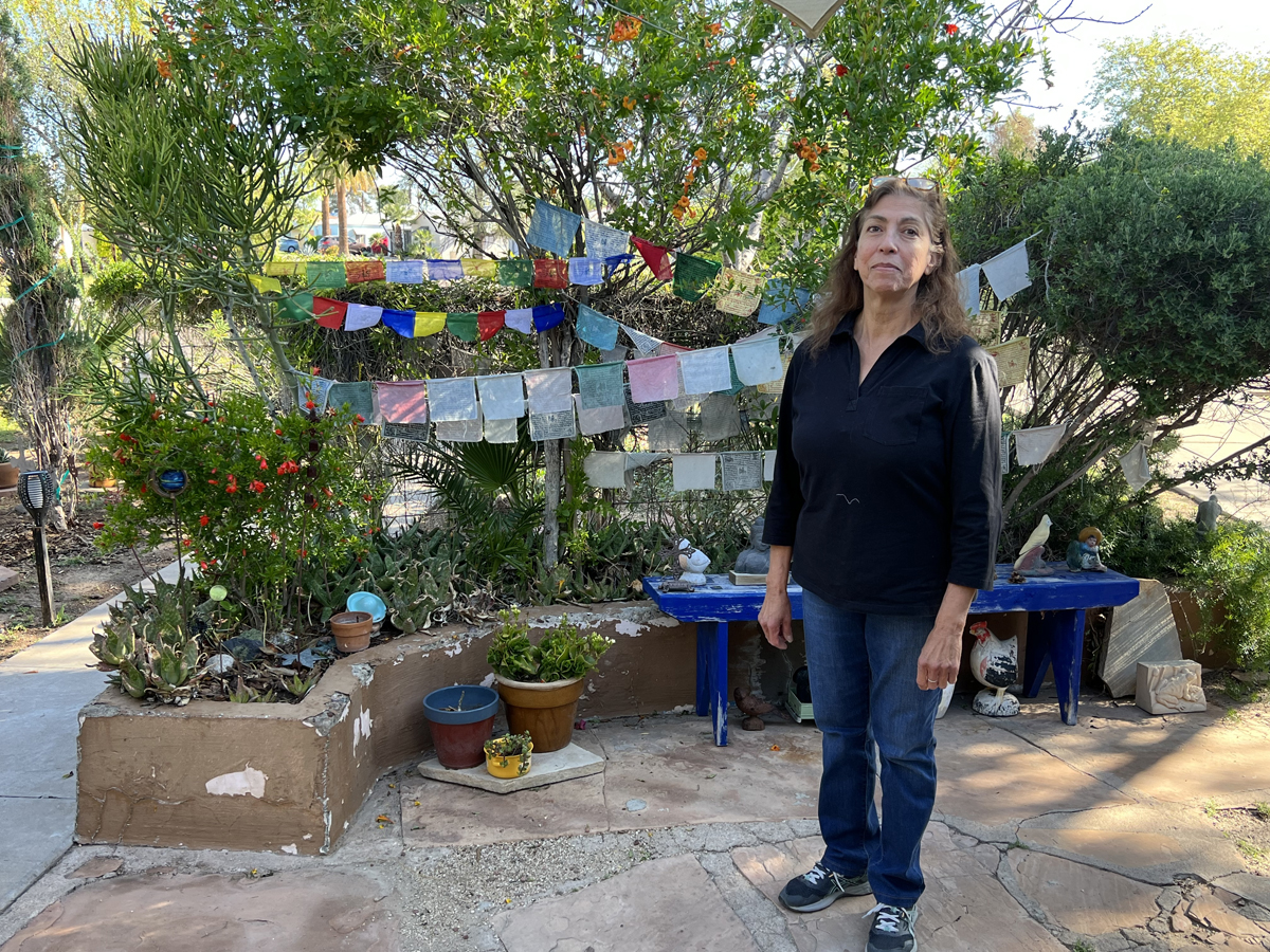 Annie Lopez stands in front of a bright blue bench in her front yard. Colorful prayer flags are strung exuberantly behind her.