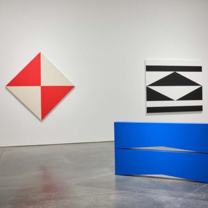 Geometric wall paintings in red, white, and black by Carmen Herrera, with a blue sculpture in the foreground.