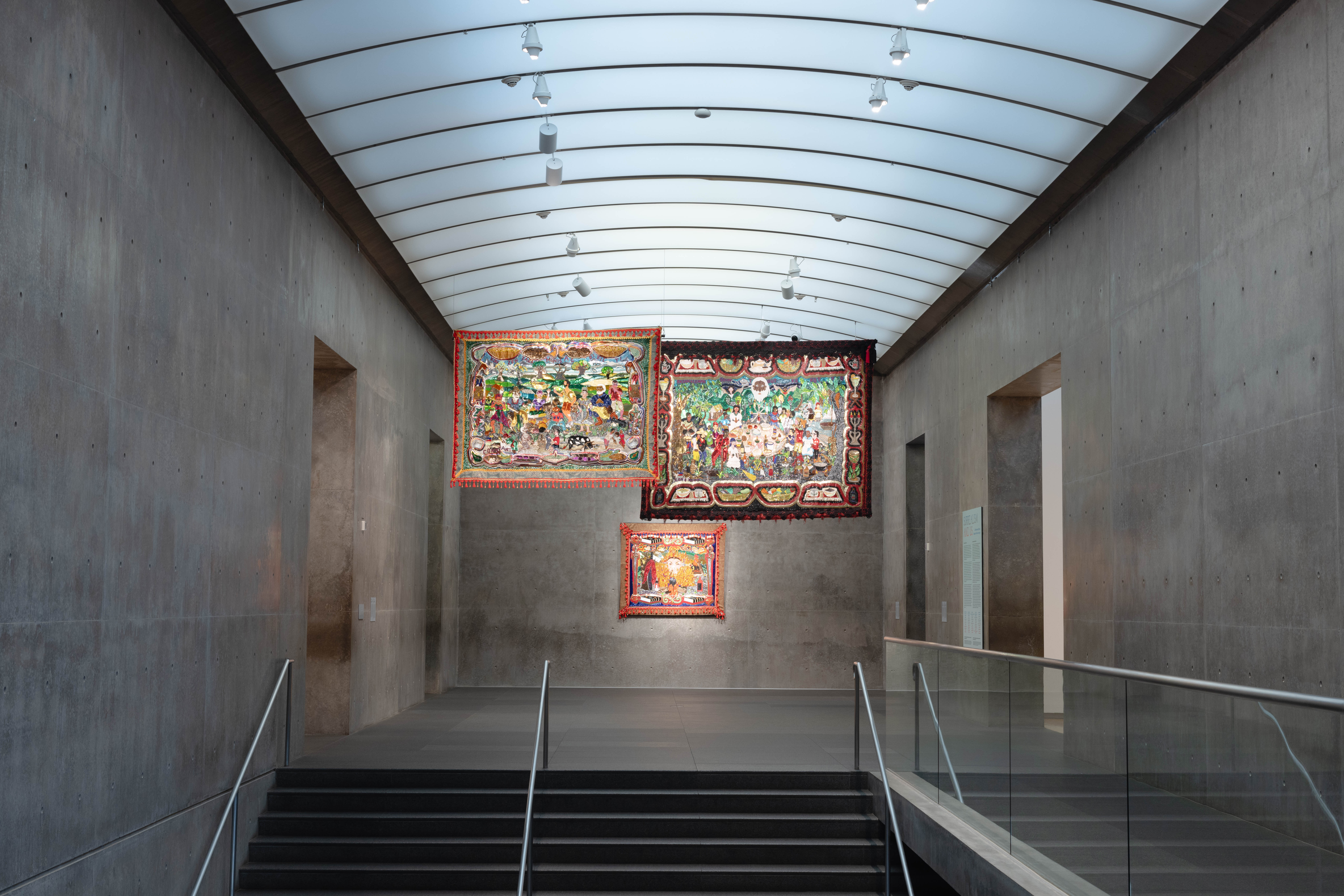 Installation view of an art space with concrete walls and skylights, with three tapestry works by Myrlande Constant suspended at center.