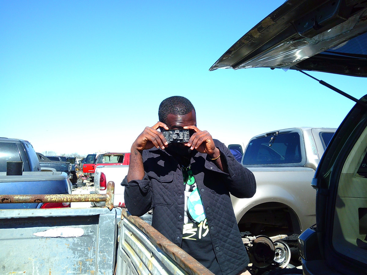 Photograph of Ghanaian artist Patrick Quarm holding a camera to his face to snap a photograph. He is surrounded by cars in a San Antonio scrapyard.