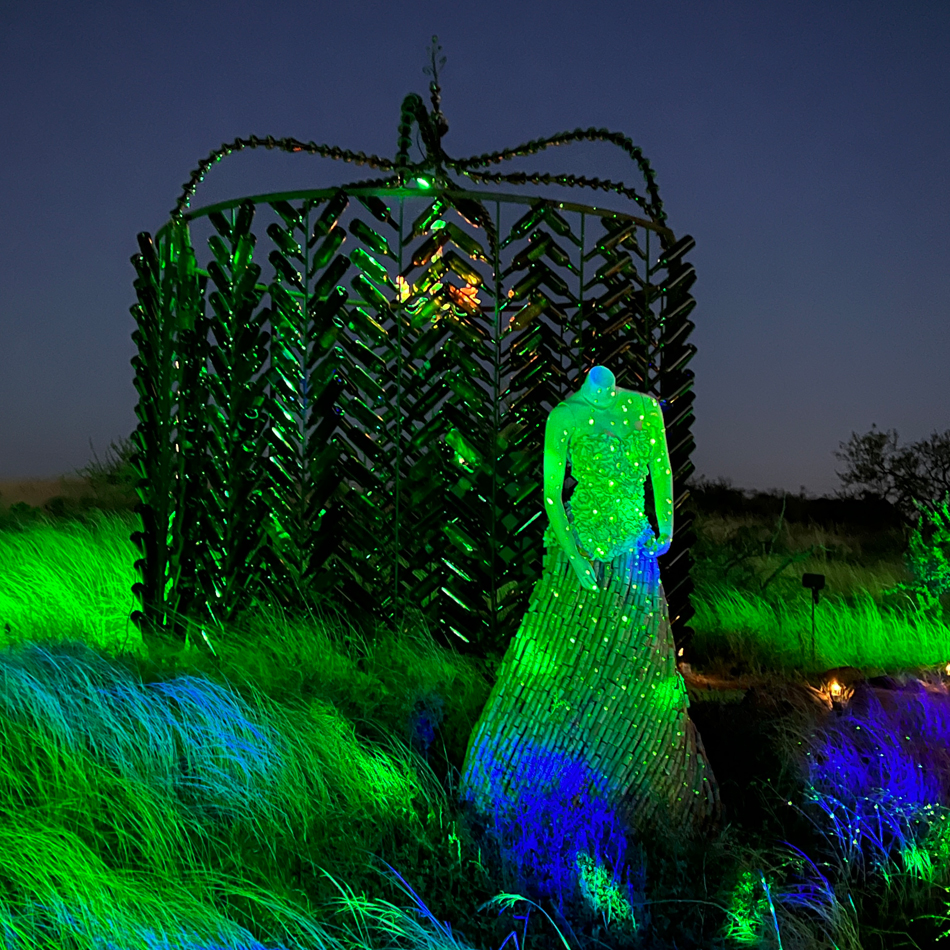 A glowing art installation set against an Arizona night sky features a glass castle and a mannequin form with a glittering dress.