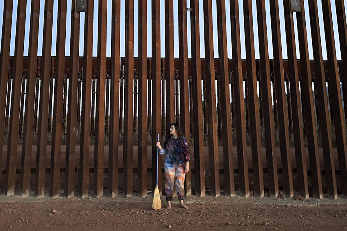 Artist Estephania González stands in front of the U.S.-Mexico border fence, a broom in hand. She is an Arizona Commission on the Arts grant recipient.