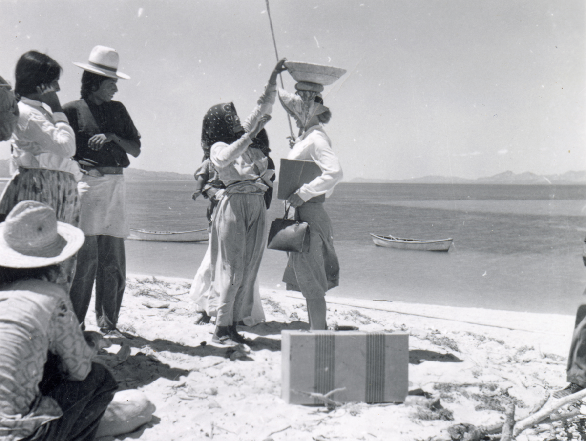 A woman balances a basket atop another woman's head while a crowd looks on. This image is featured in Woven from the Center, a new book on Native basketry by Diane Dittemore.