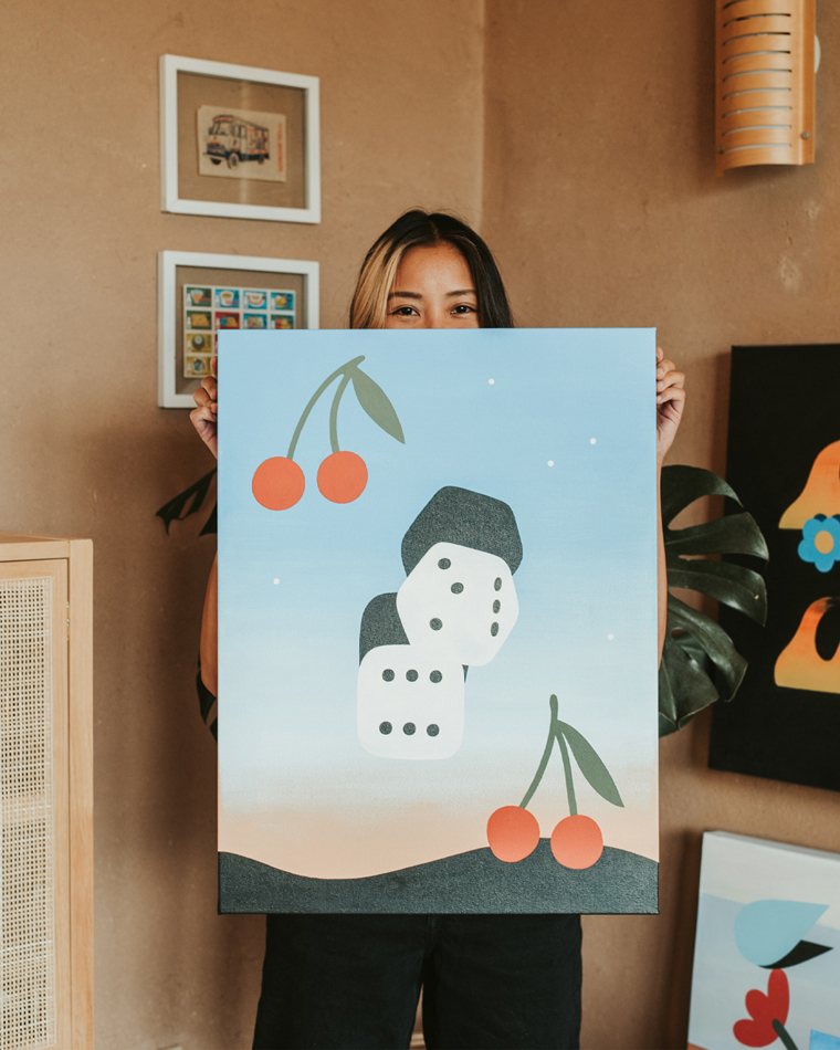 Artist and designer Paulina Ho peeks from behind a painting on canvas that depicts dice and cherries.