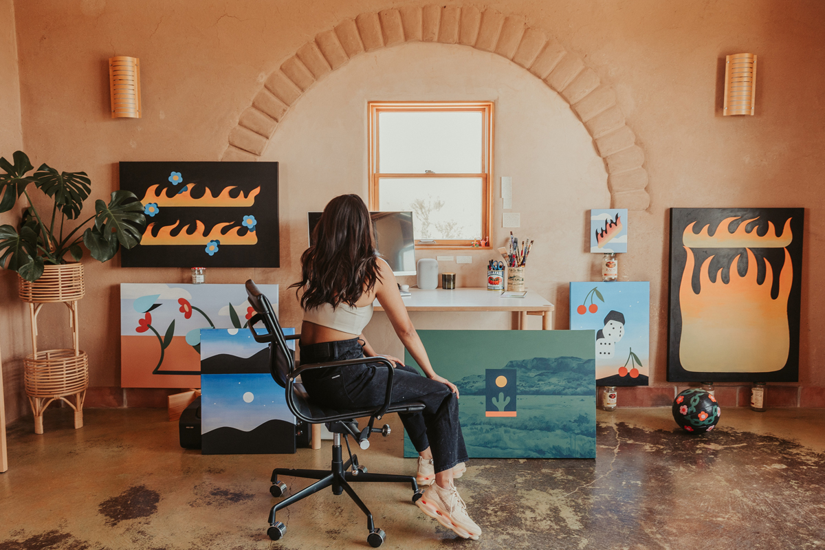 Artist and designer Paulina Ho sits in an office chair, observing a grouping of her paintings observing a grouping of her paintings in her studio in Taos, New Mexico.